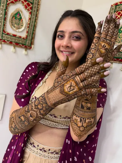 Another lovely photo of a happy bride. She is showing her full bridal mehndi, which includes personalized initials and a detailed doli scene, against a traditional backdrop.