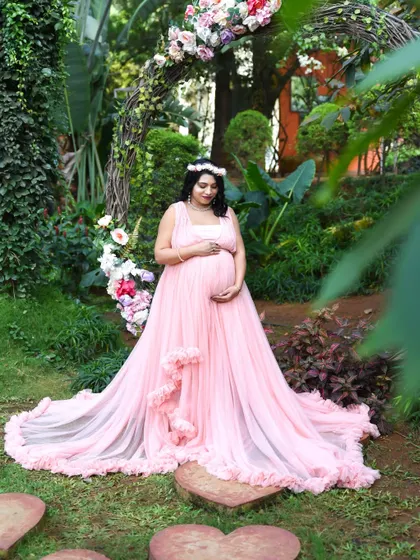 A serene moment in the garden. This mom-to-be, wearing a floral crown and a pink ruffled gown, is framed by a rustic arch and lush plants.