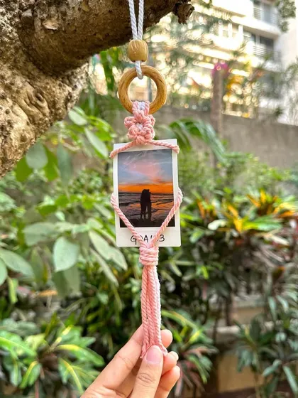 A single pink macrame polaroid holder hanging from a tree, showcasing a beautiful sunset photo. It's a perfect, personalized gift to remember a special trip or moment.