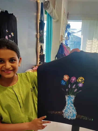 A young student smiles as she holds up her finished tote bag, which features a lovely painting of a vase of flowers. This fabric painting workshop allows kids to create functional art.