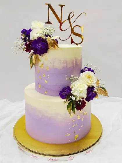 A beautiful two-tiered cake with a purple watercolor effect. It's adorned with fresh white and purple flowers, gold leaf, and a custom initial topper, making it perfect for an engagement or anniversary.