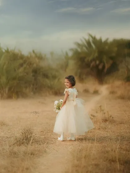 A girl in a formal dress looks back over her shoulder while walking down a path in a rustic, grassy field.