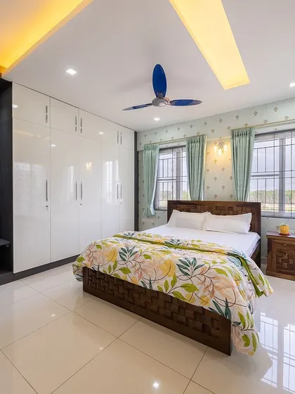 A bright and airy bedroom with white high-gloss wardrobes, a dark wood bed, and floral-patterned bedding. The large windows let in plenty of natural light.