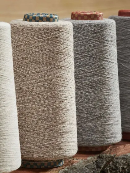A close-up shot of four large spools of yarn in neutral, earthy tones. The tight crop emphasizes the rich texture and natural feel of the threads.