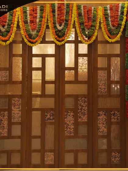 A foldable wooden door for a puja space, featuring a geometric pattern and frosted glass panels. The festive marigold toran adds a splash of color and tradition.
