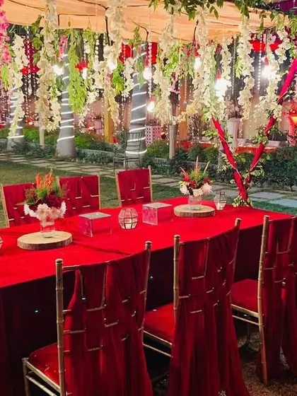 A royal red-themed seating arrangement for guests. The long table is covered in red velvet, with matching chair drapes and a canopy of hanging flowers above.