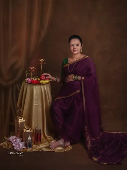 A classic portrait of a mother-to-be in a deep purple saree, styled in a traditional way. She stands next to a table with fruits and brass lamps, evoking a sense of timeless grace.