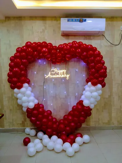 A heart-shaped balloon arch for an anniversary. The frame is made of red and white balloons with a neon 'Happy Anniversary' sign and fairy lights in the center.