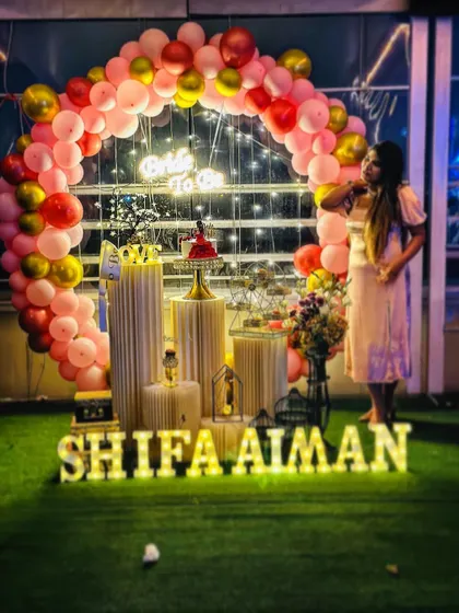 A beautiful 'Bride to Be' party setup on a terrace. The decoration includes a pink and gold balloon arch, a neon sign, and a full display of props on an artificial grass surface.