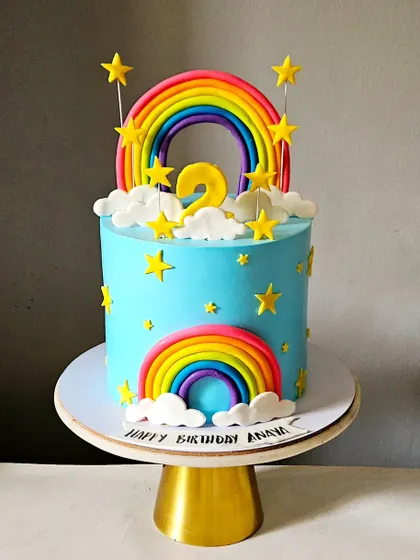 A bright and cheerful rainbow-themed cake. This tall, single-tier cake features two vibrant fondant rainbows, fluffy clouds, and scattered stars against a brilliant blue sky.