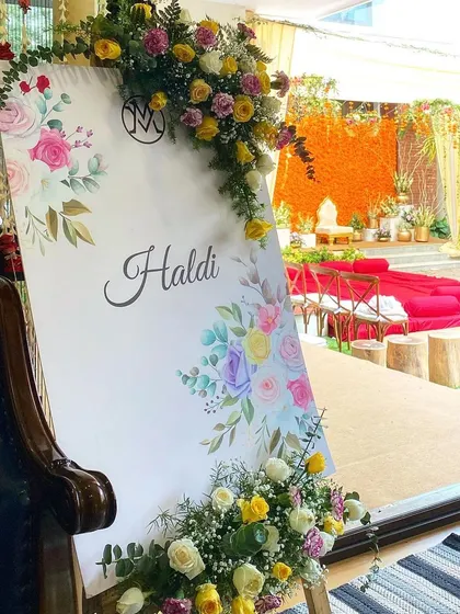 A pretty welcome sign for a Haldi ceremony, adorned with fresh yellow roses and baby's breath. In the background, you can see the main event space with its bright marigold decor, setting the tone for a fun-filled event.