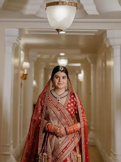 A full-length portrait of the bride in the hallway of The Leela Palace, showcasing her stunning red and beige lehenga.