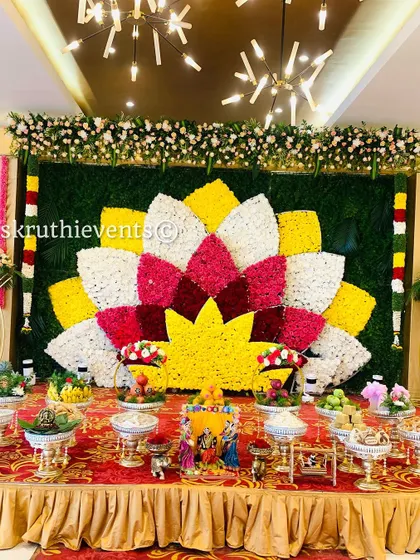 A beautiful offering display set before a large floral lotus mosaic. The arrangement includes traditional dolls and a variety of sweets and fruits presented in decorative bowls.