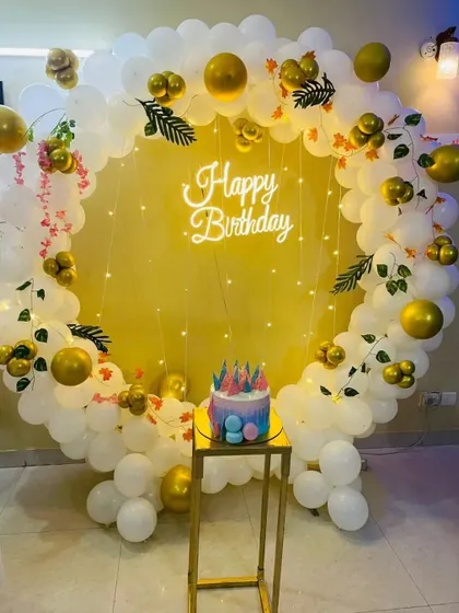 A simple and elegant birthday photo corner with a circular balloon arch in white and gold, a neon sign, and a plinth for the cake.