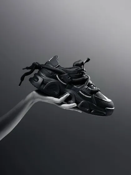 A dramatic black and white shot of a hand holding a chunky black sneaker. The minimalist composition and high-contrast lighting give the product a powerful and iconic look.