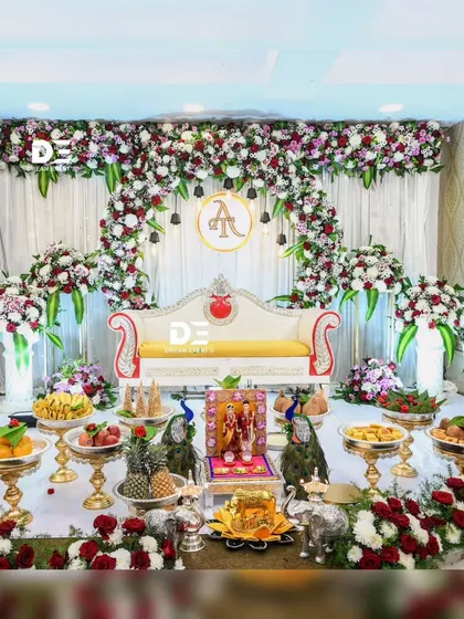 This traditional engagement ceremony setup is rich with detail. The floral arch is complemented by a display of fruits, sweets, and traditional dolls, creating a scene that is both beautiful and culturally significant.