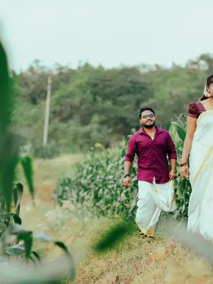 A couple walks through a field of tall green plants, dressed in traditional attire. The shot captures a sense of journey and togetherness.