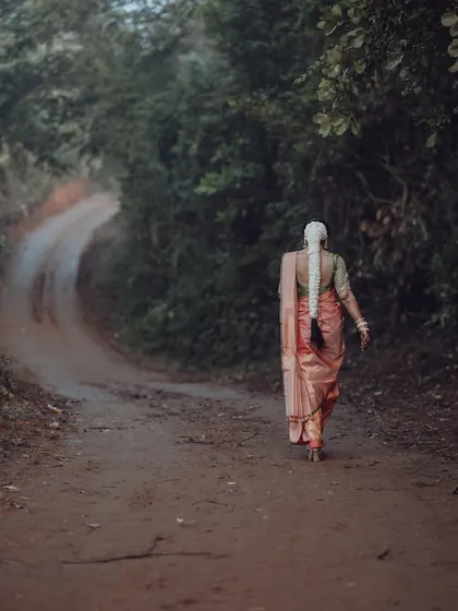 A South Indian bride walks down a rustic path, her traditional saree and long braided hair creating a picture of timeless elegance.