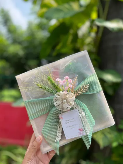 A softly wrapped gift in a pastel green theme, held in hand to show its delicate details, including a lace-edged ribbon and a custom tag.