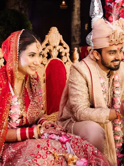 The newlyweds seated in the mandap, sharing a happy moment. The ornate golden chairs and the rich red floral backdrop create a regal frame for the couple.