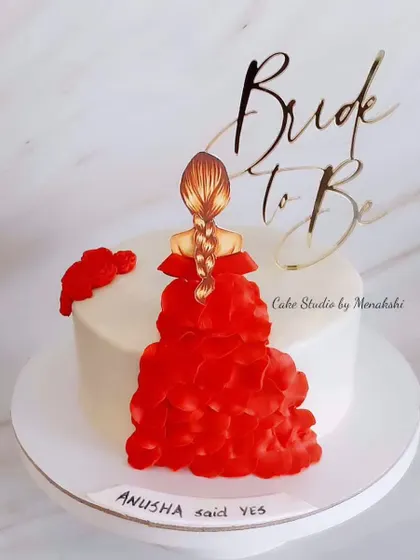 A pretty cake for a bride-to-be. This elegant design features a topper of a bride in a beautiful red dress, celebrating her upcoming wedding.