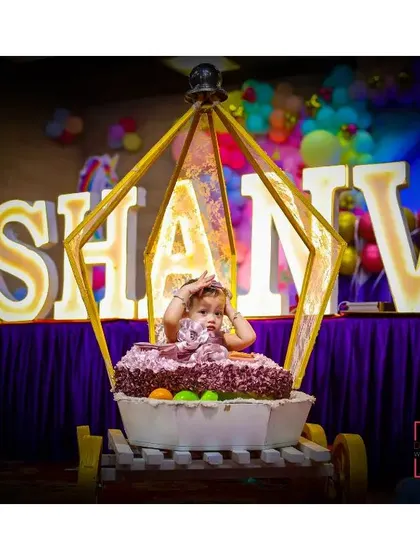 A grand first birthday setup with the birthday girl, Shanvi, sitting in a custom carriage-like high chair in front of her name in large, lit-up letters.