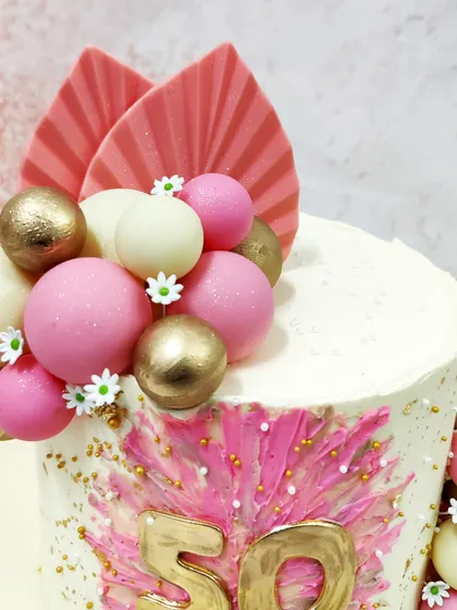 We celebrate a 50th birthday with this vibrant and modern pink boho cake. A bold pink starburst design with a gold "50" is the centerpiece, complemented by pink, white, and gold spheres and dried palm leaves for a chic, bohemian look.