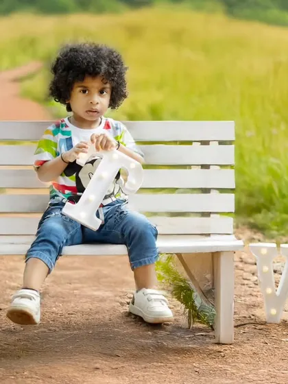 A toddler boy with amazing curly hair sits on a bench during his outdoor second birthday shoot.