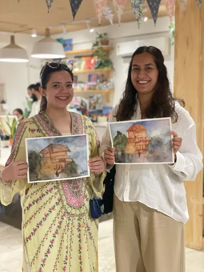 Two friends proudly hold up their watercolor landscape paintings. Our mentors guide you through the process, so you can create something you're truly happy with.