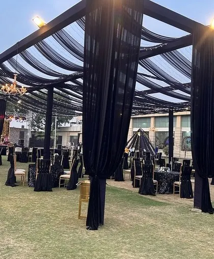 A large-scale outdoor setup for a black-themed cocktail event. The decor includes black drapes, black table linens, and elegant chandeliers.