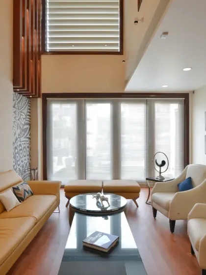 The first-floor living room of the Gupta Residence is a modern, inviting space with contemporary seating and wooden flooring. Large windows fill the room with natural light, while backlit shelving and framed photographs add personal character.