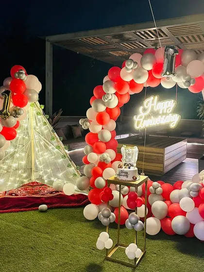 A beautiful first-anniversary celebration on a terrace. This setup features a red and white balloon arch, a neon sign, and a cozy canopy tent with fairy lights.