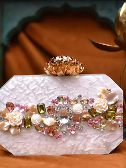 A beautiful white acrylic clutch featuring a mix of colorful rhinestones and delicate floral details. The lotus-shaped clasp adds a unique and luxurious finishing touch.