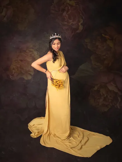 A portrait of a queen awaiting her little one. The mom-to-be wears a crown and a lovely yellow gown, holding a small bouquet against a rich, floral backdrop.
