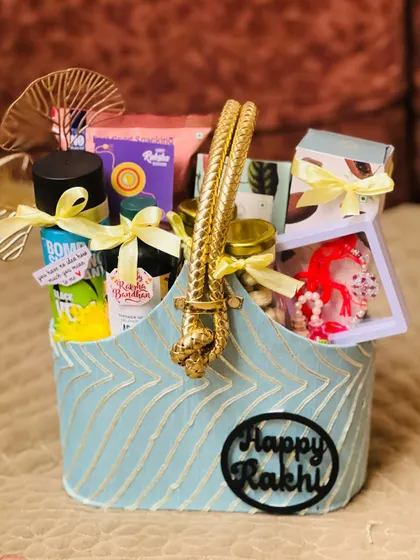A chic blue fabric basket with a thick gold rope handle, filled with Rakhi gifts and a "Happy Rakhi" tag.