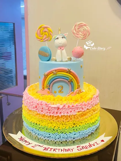 A two-tier unicorn cake with a rainbow-colored ruffle design on the bottom tier. The cake is topped with a fondant unicorn, lollipops, and a rainbow.