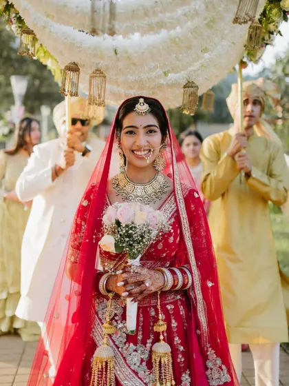 The bride makes her grand entrance, smiling brightly as she walks under a beautiful phoolon ki chadar held by her family.