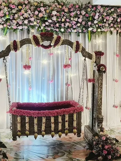 A grand and traditional cradle ceremony setup. The wooden cradle is decorated with pink flowers and placed in front of a stage draped in white curtains, all framed by an extensive arch of pink and white flowers.