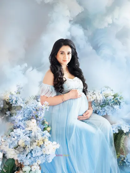 A beautiful, fantasy-themed portrait of a mother-to-be. She is seated in a light blue gown amidst a bed of blue flowers and soft, cloud-like textures, creating a serene and magical atmosphere.