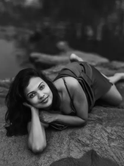 A gentle black and white portrait of a woman lying on a rock formation, looking softly at the camera. The blurred natural background focuses attention on her relaxed and serene expression.