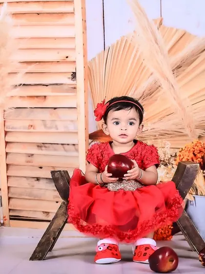 A baby girl in a red dress sitting in a rustic setup with apples and pampas grass.