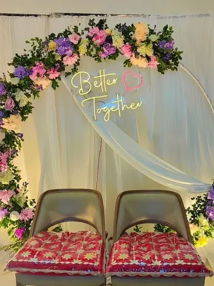 A beautiful floral ring backdrop for an intimate engagement or sangeet. The arch is densely decorated with a mix of colorful artificial flowers and features a "Better Together" neon sign, adding a romantic glow to the couple's seating area.