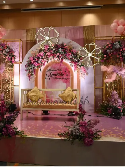 A glamorous stage setup for an Aqeeqah ceremony. The decor is a beautiful blend of pink, purple, and gold, with a floral arch, neon flower lights, and a comfortable seating area for the family.