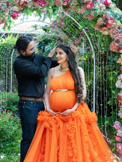 A playful and loving moment between a couple. The dad-to-be gently places a crown on his partner's head as she sits in a floral arch, both sharing a happy smile.