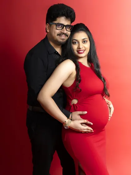 A classic studio portrait against a vibrant red background. The couple's pose is confident and happy, celebrating the new life they are bringing into the world.