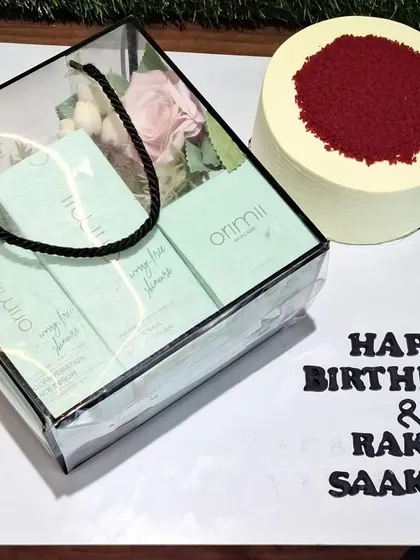 A combined celebration gift for a birthday and Rakhi. This features a clear bag hamper filled with skincare products, presented alongside a delicious red velvet cake.