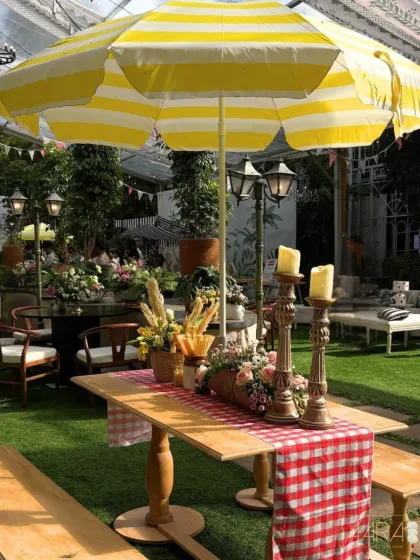 A picnic-style seating area from the 'Borough Bazaar' theme. A yellow and white striped umbrella provides shade over a wooden table decorated with a gingham runner and fresh flowers in a bread basket.