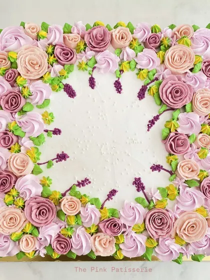 A square cake that serves as a canvas for intricate floral piping. The border is a lush garden of buttercream roses and leaves in shades of pink, purple, and lavender.