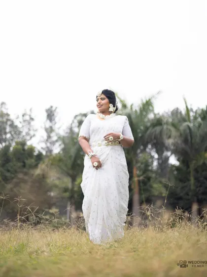 A beautiful portrait of the bride in a white saree and floral jewelry, standing in a serene outdoor setting for her Haldi ceremony.
