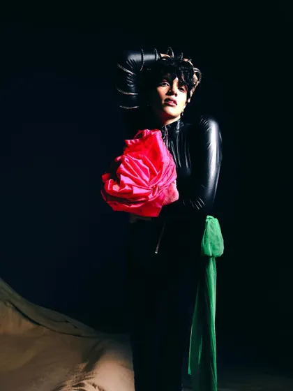 A full-body shot with the model set against a dark background, holding a vibrant pink floral piece. The lighting creates a spotlight effect, drawing all attention to the subject and the pop of color.